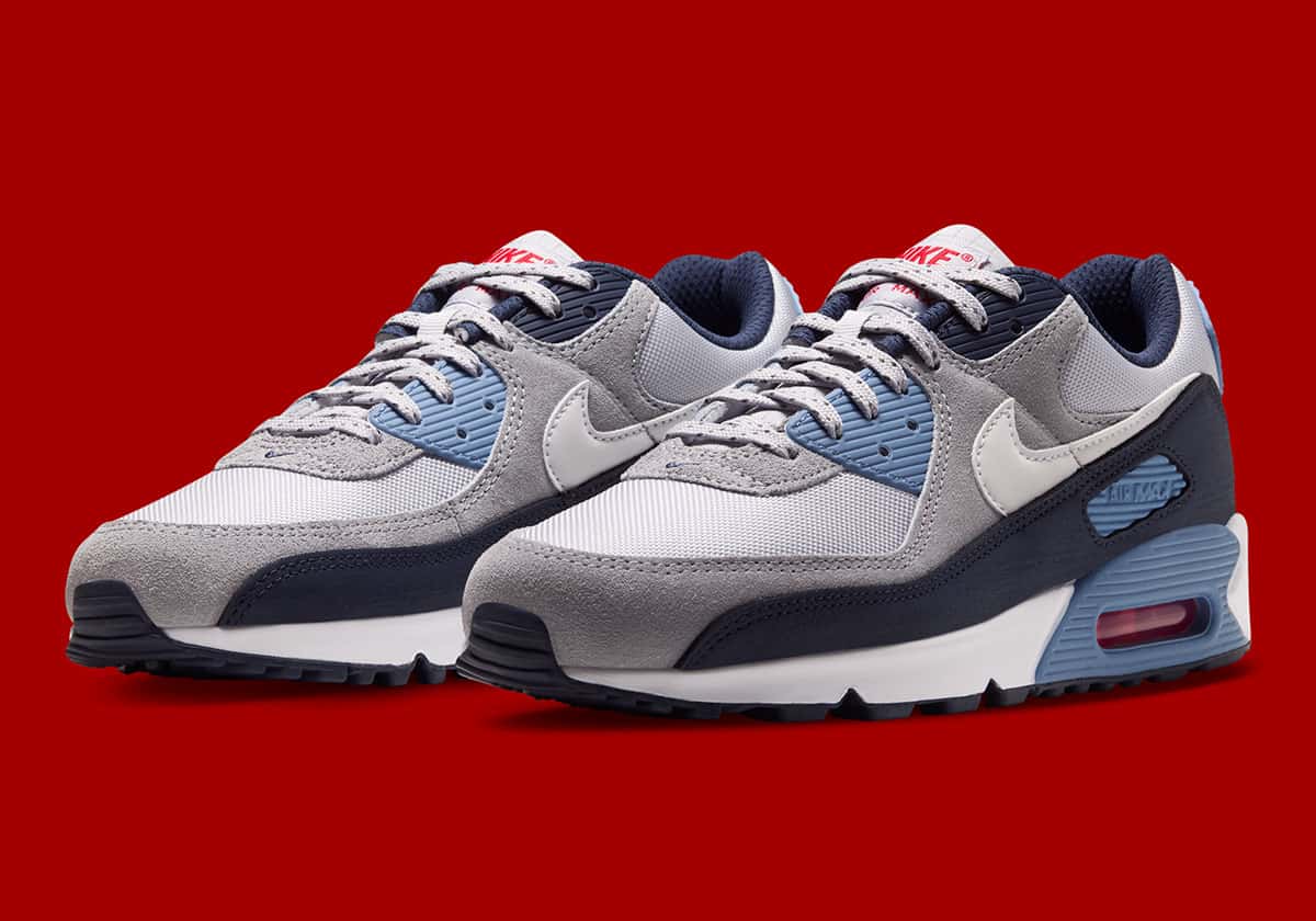 Par de Nike Air Max 90 Geyser Grey Work Blue en diagonal sobre fondo rojo, con malla gris clara, gamuza gris, Swoosh blanco, puntera azul marino, detalles Work Blue en la mediasuela y aire rojo a la vista