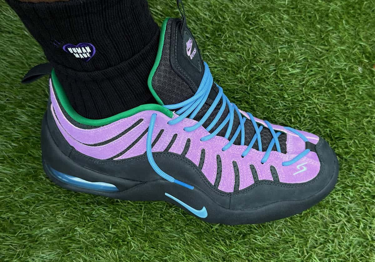 Jae Tips x Nike Air Bakin Low Purple Stardust puesto sobre pasto, con overlays de suede violeta, cordones celestes, cuello verde, Swoosh celeste y base negra